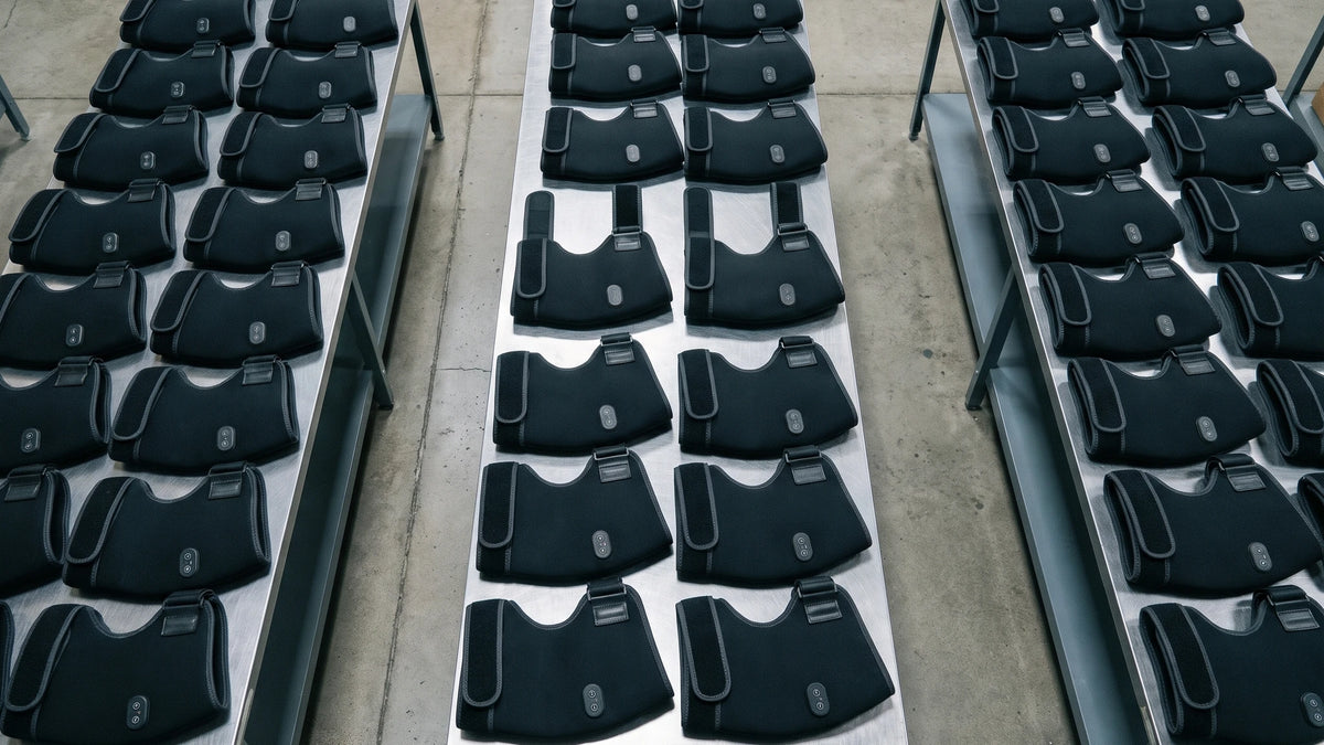 Vus d'en haut, trois longues tables d'entrepôt couvertes de dizaines de masseurs de hanche 4-en-1 noirs, posés à plat côte à côte